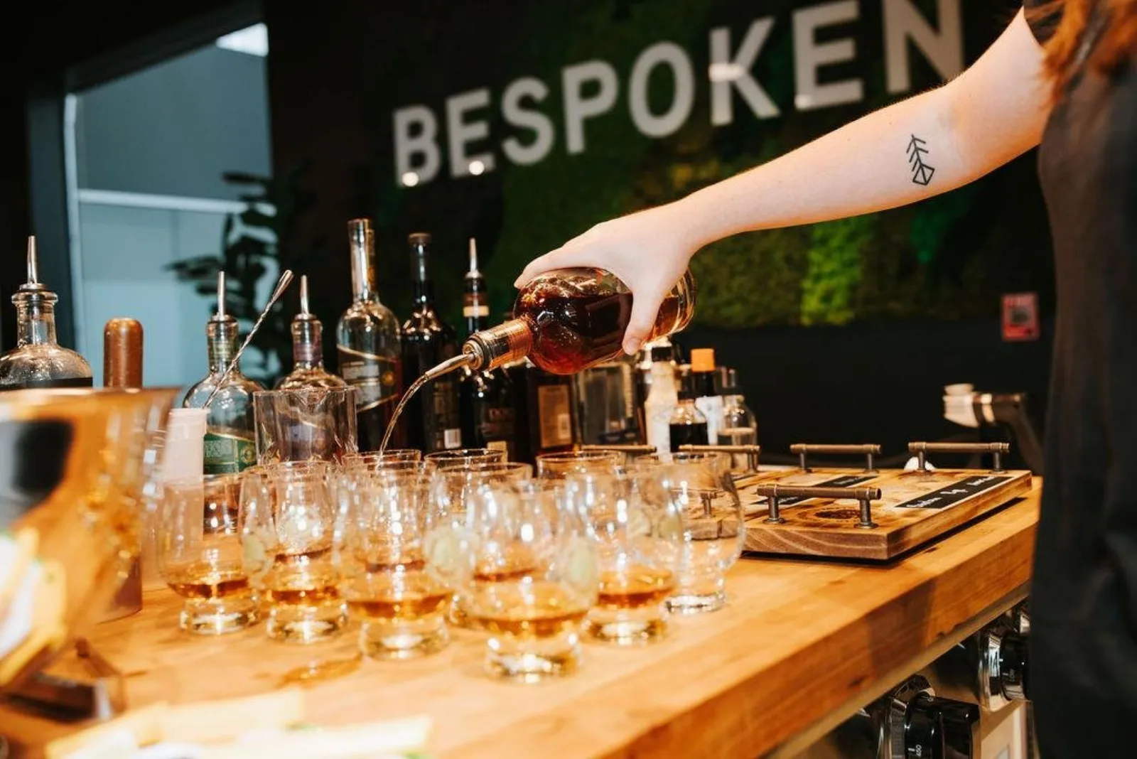 A tattooed bartender pouring bourbon into a row of tasting glasses at a Lexington distillery